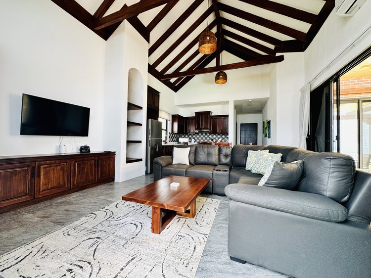 Living room with vaulted wooden ceiling, gray sectional sofa with pillows, wooden coffee table, wall-mounted TV, and open kitchen in background with dark cabinetry and patterned backsplash.